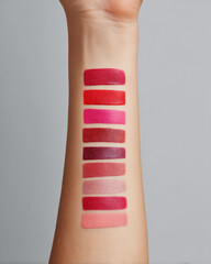 Hand displaying variety of lipstick swatches aligned vertically on forearm, showcasing spectrum of colors from deep reds to soft pinks, creating artistic and vibrant visual appeal