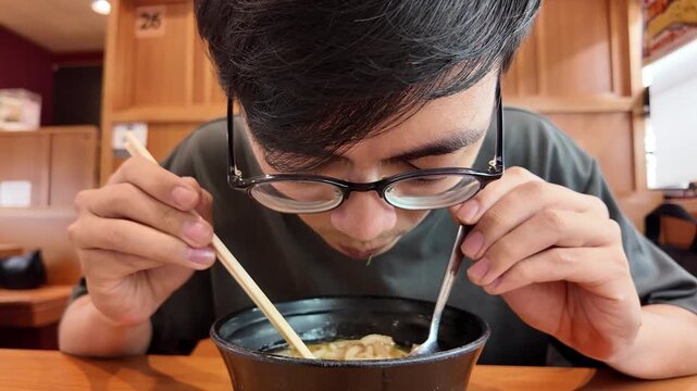 A man with glasses heartily slurps noodles from a bowl of ramen, using chopsticks and a spoon, showcasing the authentic Japanese dining experience.