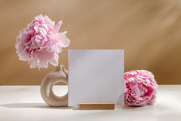 Square mockup paper card is standing on wooden paper holder with peony flowers on brown background...