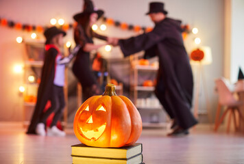 Glowing carved pumpkin standing on books in decorated room while blurred family in Halloween...