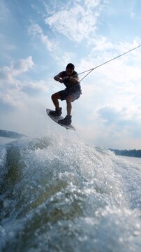 Wakeboarder performing a jump, holding the rope and balancing on the board over the water surface, creating a splash against a backdrop of cloudy sky
