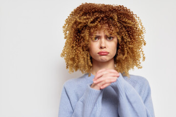 Sad young woman with curly hair expressing concern in a light blue sweater against a plain white...