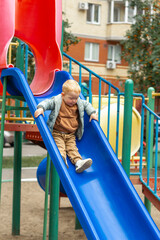 A little boy in a sweatshirt and jacket slides down a bright blue slide on a playground in the...