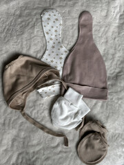 Beige and white hats with stars, a bonnet, and scratches lie on the light blanket of the bed in the room. A set of accessories for a newborn baby. Top view