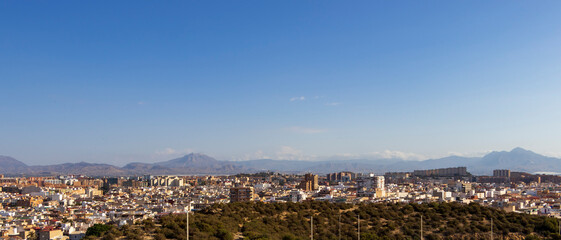 Obraz premium A panoramic view of Alicante city showcasing modern buildings and historical architecture under a clear blue sky. The vibrant landscape highlights the natural beauty of the surrounding mountains.