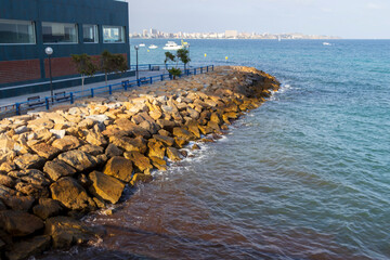 Seaside view of a rocky promenade at Alicante, Spain, where waves gently lap against the stones. A...