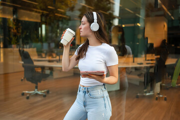 Young woman drinking coffee and using tablet wearing headphones outside office