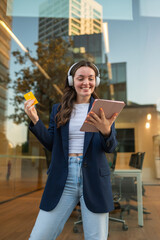 Young businesswoman buying online using tablet and credit card, wearing headphones and smiling near office building
