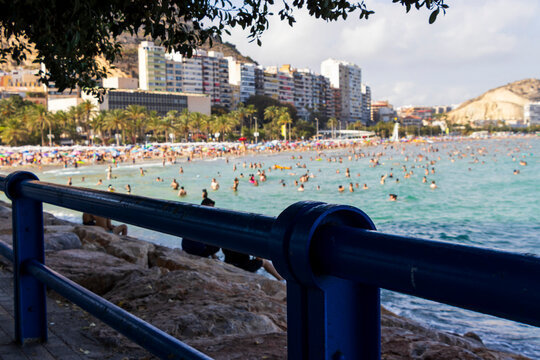 Vibrant beach scene at Playa del Postiguet in Alicante featuring a crowded shoreline with sunbathers and palm trees, showcasing summer enjoyment and leisure activities.