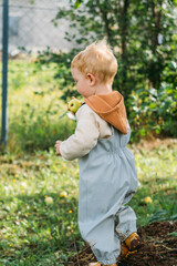 A small red-haired boy in a gray-blue jumpsuit and yellow boots holds an apple in his hands under an apple tree