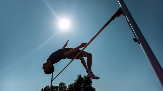 High jumper silhouetted against blue sky during athletic competition in bright sunlight. Concept of training documentation, practice vaults, sports skill refinement