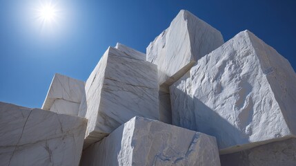 Large, white marble blocks are stacked high in a quarry, illuminated by the bright sun against a clear blue sky, showcasing the beauty and potential of raw marble