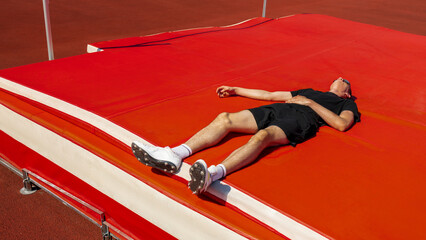 Athlete resting on red high jump mat after intense training session in athletic field. Concept of...
