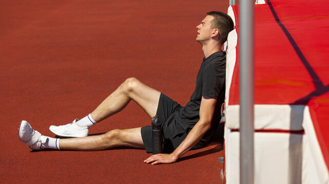 Athlete resting on track field under bright sunlight against high jump mat. Concept of physical recovery, post-jump fatigue, athletic exhaustion and mental reset