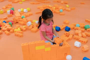 A young girl with pigtails in a pink shirt playing with large orange building blocks on an orange floor.