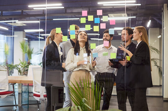 Group of professionals working in modern office, using colorful sticky notes on glass board for brainstorming and collaborative planning. Company employees discussing business project during teamwork.