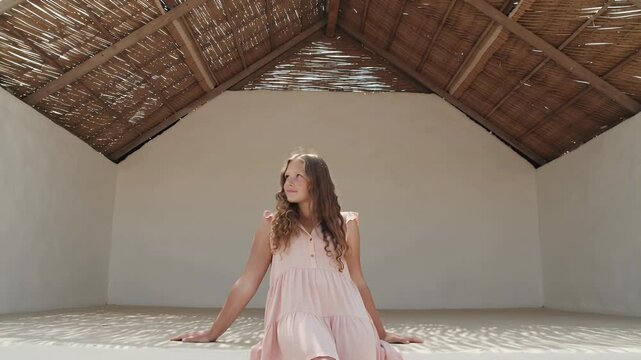 A girl in a light pink dress sits beneath a thatched roof