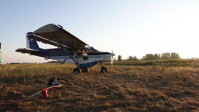 agricultural aircraft parked on dry rural airfield during golden hour, liquid transfer pump connected to underwing sprayer, peaceful countryside, evening, agrotechnology, aviation, farming