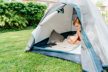 School-age boy sitting in his tent outdoors and reading a book. Summer holidays.
