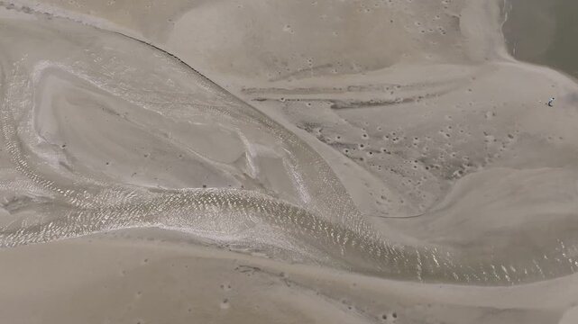 A winding water channel glistens in the sun, snaking through soft, pale sands with intricate ripples and tide-carved impressions.