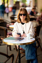 Middle aged woman sitting at sidewalk cafe in the city and using smartphone