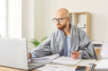 Sick office employee man wrapped in warm blanket working at laptop suffering from cold, fever, flu,...