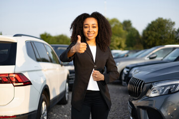 A happy businesswoman stands in front of cars giving a thumbs up, possibly selling a vehicle at a...