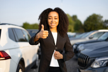 A woman, car salesperson, gives a thumbs-up, indicating approval, at a car dealership. Cars are...