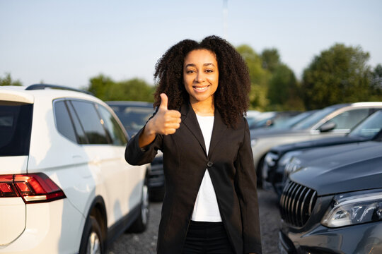 A smiling woman giving a thumbs up in a car lot, suggesting a positive car-buying experience.