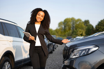 Smiling car salesperson presenting a vehicle on a sunny day. The woman holds a tablet and gestures...