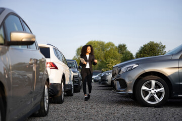 A businesswoman walks through a car lot, possibly selling vehicles. She is looking at the cars, possibly for a customer.