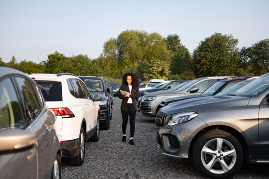 A businesswoman reviews paperwork between rows of cars in a dealership lot, assessing vehicle options.