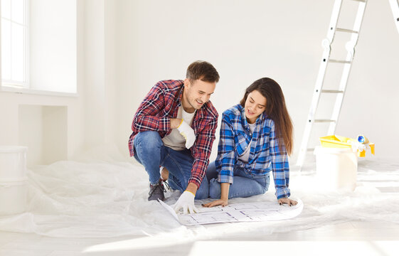Fototapeta Happy young couple sitting on floor with blueprint in new apartment and discussing design project of their house. Man and woman planning repair in new flat. Renovation and moving concept.