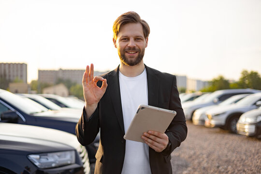 A car salesman smiles while holding a tablet, making an 'okay' gesture, with cars in the background, perfect for automotive advertising. - Powered by Adobe