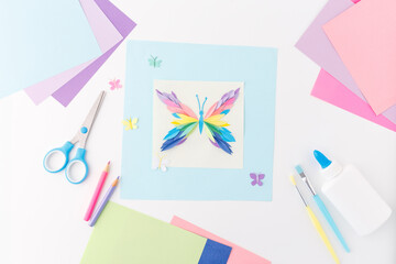 Colorful butterfly made from layered paper cutouts surrounded by craft tools and colored sheets. Flat lay composition with copy space. Paper art and kids' craft concept. Design