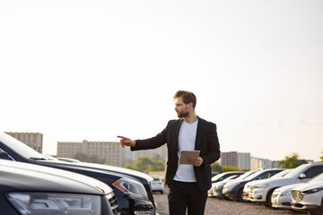 A businessman in a suit points towards a car in a parking lot, holding a tablet while reviewing the...