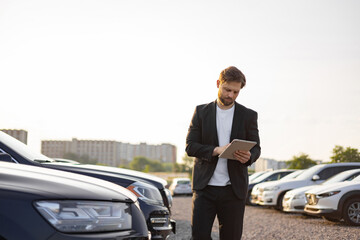 A businessman in a suit reviews a tablet while standing near parked cars in a car dealership.