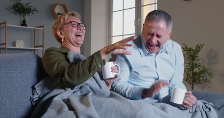 Happy elderly married couple spending an evening at home living room, laughing while watching a comedy movie on television. Celebrating family moments of leisure time and shared joy. - Powered by Adobe