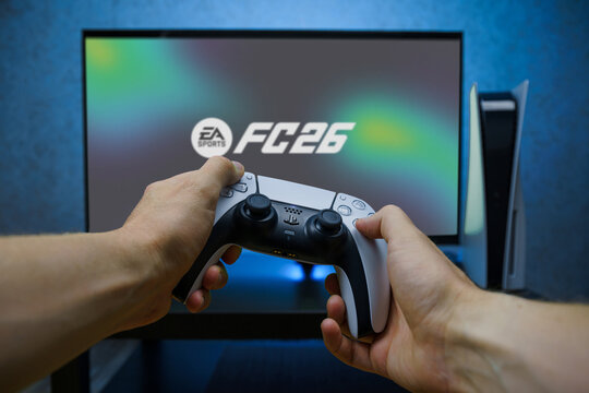 Hands holding a PlayStation controller in front of a screen showing EA Sports FC 26 on a gaming console.