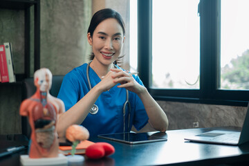 Female doctor in blue scrubs sitting at desk with medical models and tablet, smiling confidently in...