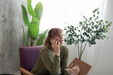 Woman talking on phone and holding a book while sitting in armchair