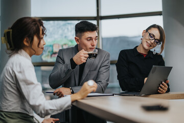 Three business people are in a relaxed meeting, with one sipping coffee while discussing work on a...