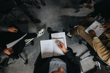 Business people are seated in a circle, engaging in a meeting. Papers and notebooks are visible, indicating active participation and discussion. Overhead view emphasizes teamwork and communication.