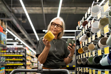 Stylish mature woman shopping for home goods in a modern retail store selects decor and kitchen...