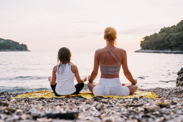 Mother and daughter practicing yoga at seaside at sunset or sunrise.