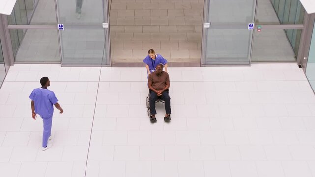 Overhead wide shot of female nurse wearing scrubs pushing senior man in wheelchair across lobby of modern hospital - shot in slow motion