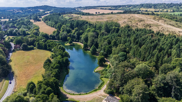 Aerial view of a serene lake mirroring the sky, framed by dense green trees and open fields under a bright sky, Guildford, England, United Kingdom.
