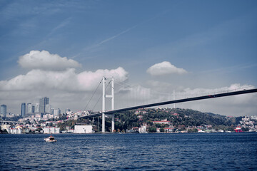 Naklejka premium A magnificent suspension bridge connecting a bustling city with modern architecture across calm waters, featuring lush green hills, a boat on the water, and a serene cloudy blue sky.