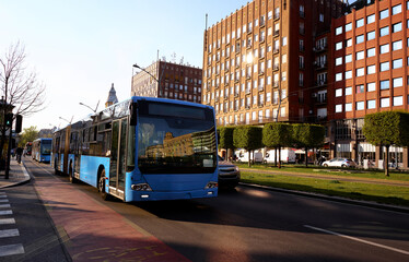 Obraz premium Modern Blue City Bus Driving Through Urban Street Amidst Evening Sunlight