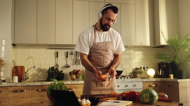  Brutal man cooking in the kitchen, peeling vegetables while watching a cooking tutorial on a tablet in headphones. Nutrition, healthy lifestyle, technology, mindfulness, relax, learning, tablet